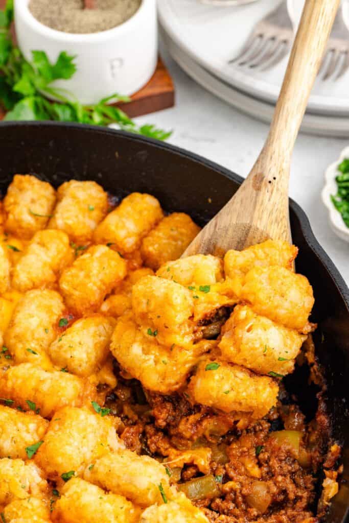 Wooden spoon lifts cheesy sloppy joe tater tot casserole with ground beef from a cast iron skillet.