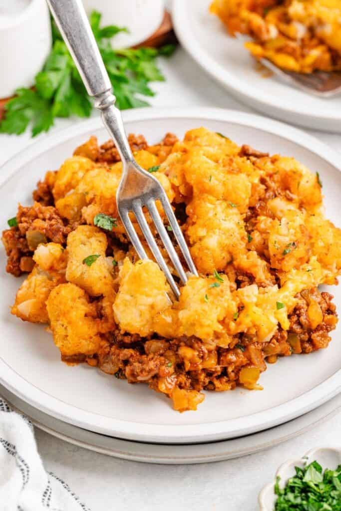 Sloppy joe tater tot casserole with ground beef, cheese, and vegetables on a white plate, fork resting on top.