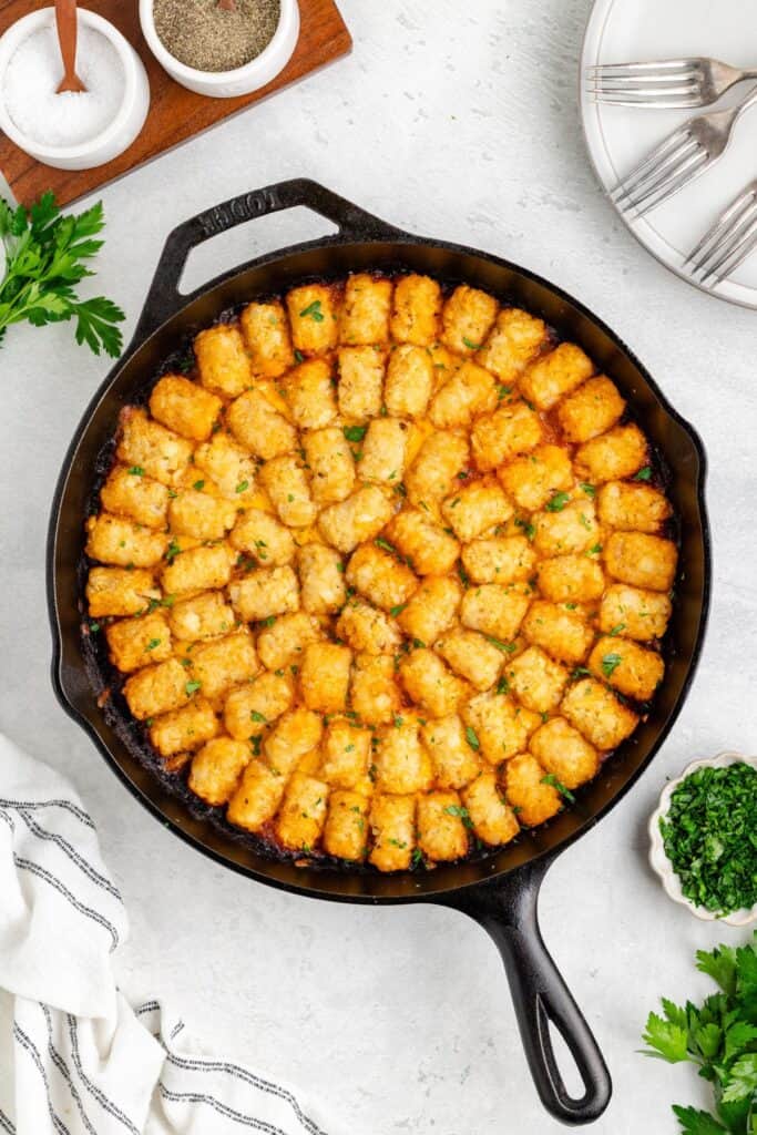 Cast iron skillet with tater tots sloppy joe casserole garnished with herbs, surrounded by plates, utensils, and seasonings.