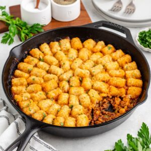 Cast iron skillet of sloppy joe tater tot casserole with golden tots, ground beef, vegetables, and parsley garnish.
