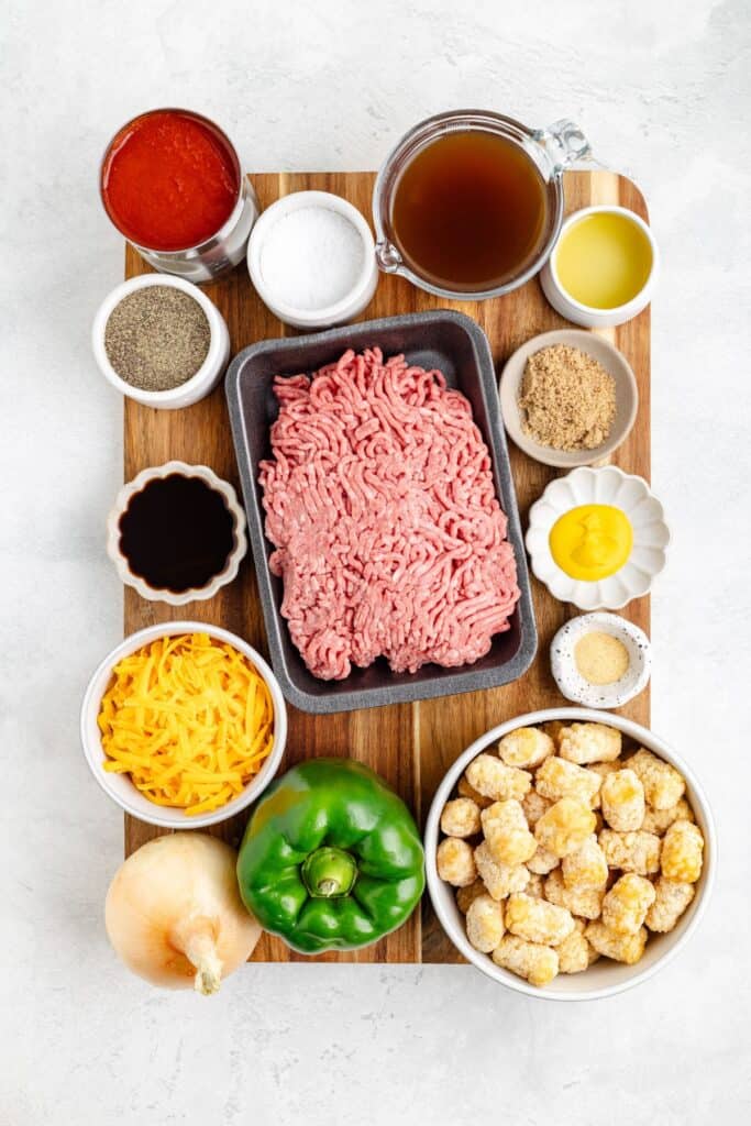 Ingredients for a recipe arranged on a wooden board: ground beef, tater tots, vegetables, cheese, and various seasonings.