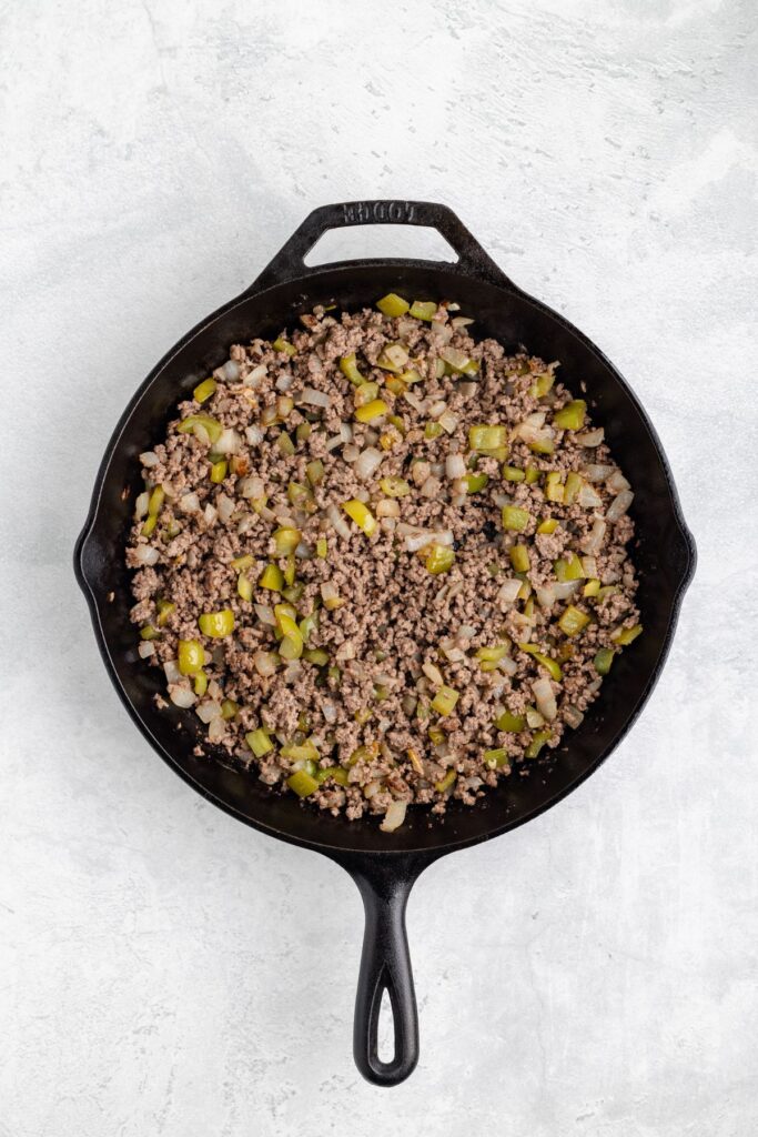 Cast iron skillet with cooked ground beef, diced onions, and green bell peppers on a light gray surface.