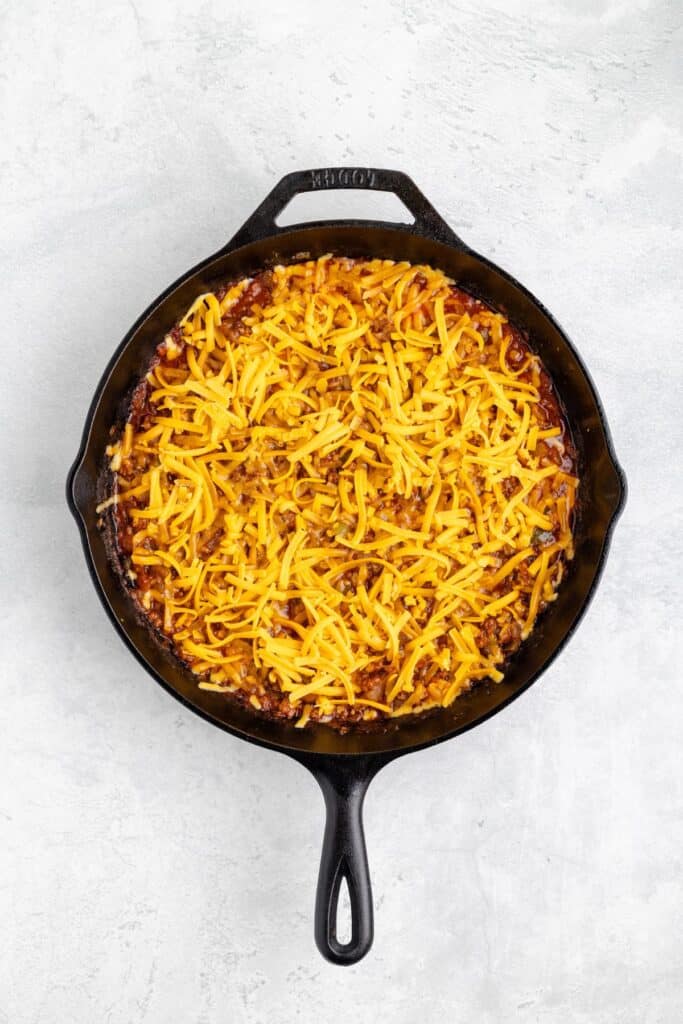 Cast iron skillet with a sloppy joe sauce topped with melted cheddar cheese on a light gray surface.