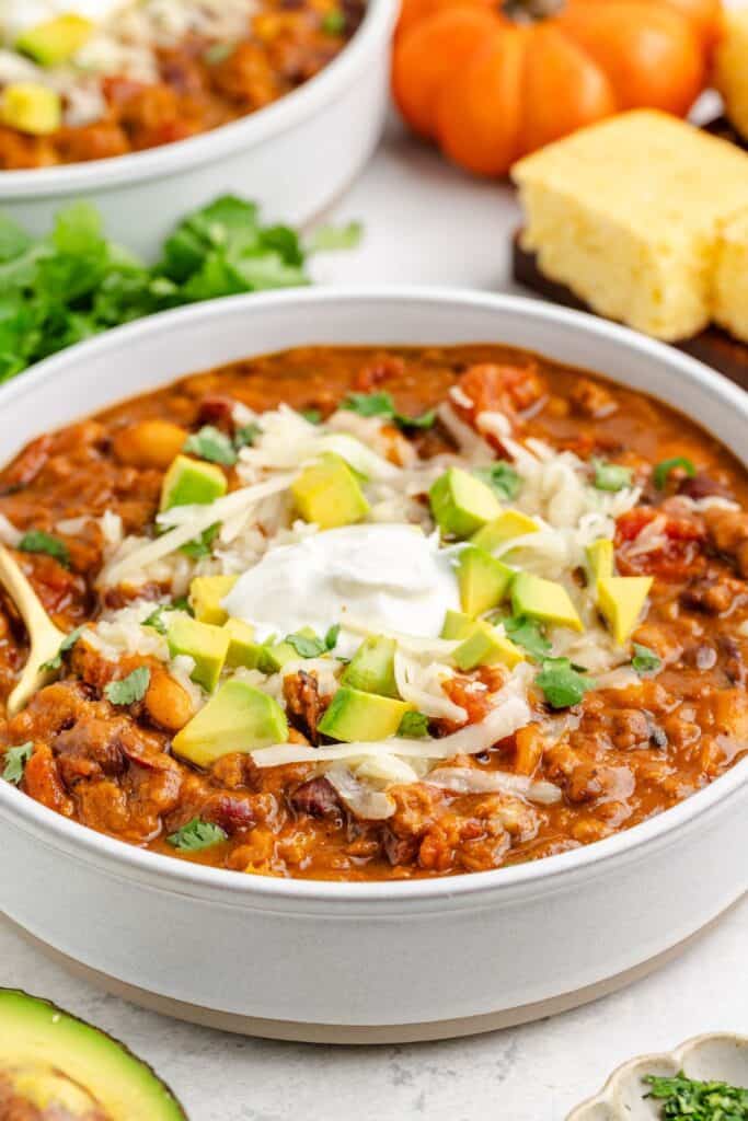 Pumpkin turkey chili in a bowl topped with sour cream, avocado, cheese, and cilantro; cornbread and veggies behind.