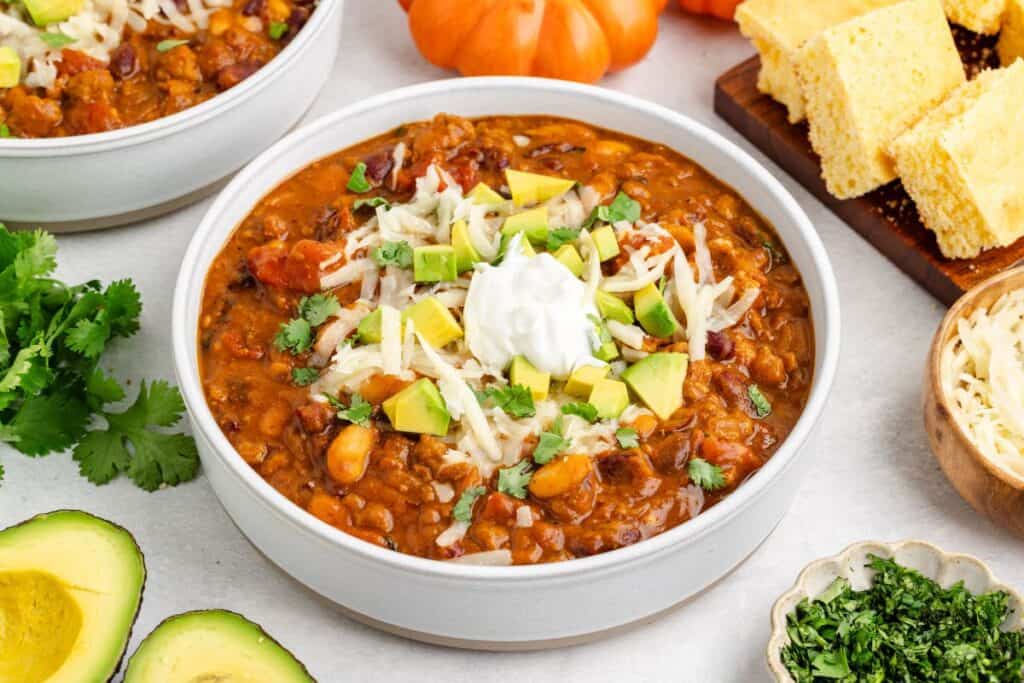 Bowl of turkey pumpkin chili with avocado, shredded cheese, and sour cream, surrounded by cilantro, avocado, cornbread, and cheese.