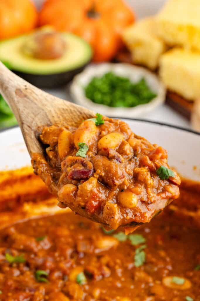 A wooden spoon holds chunky pumpkin chili with beans and ground turkey over a pot, with cornbread and avocado behind.