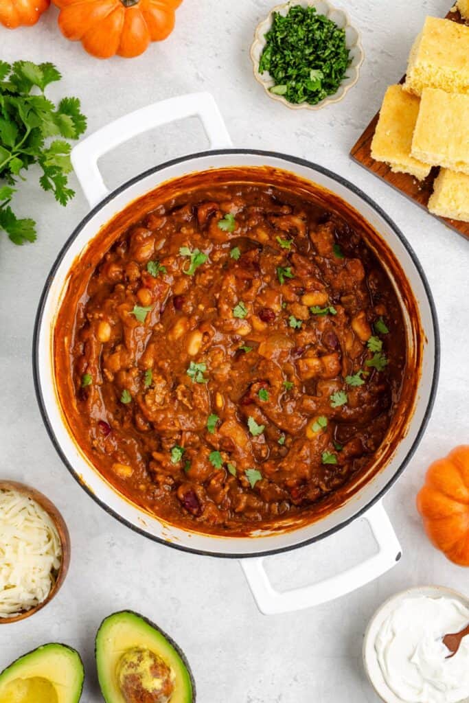 Pot of pumpkin chili with beans and vegetables, topped with herbs, surrounded by cornbread and assorted toppings.