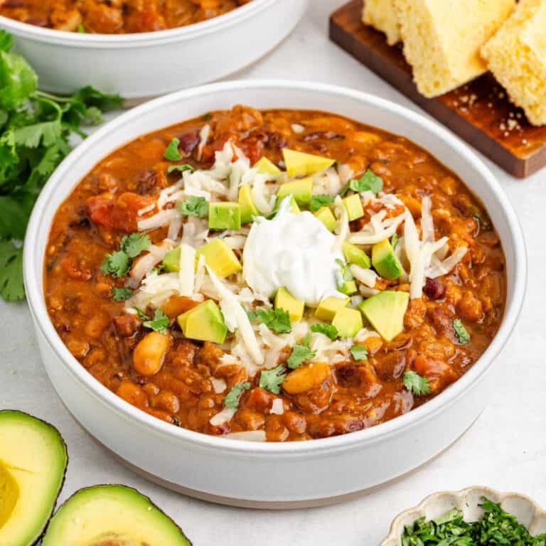 Bowl of pumpkin chili with turkey topped with sour cream, cheese, avocado, cilantro; cornbread and herbs on the side.