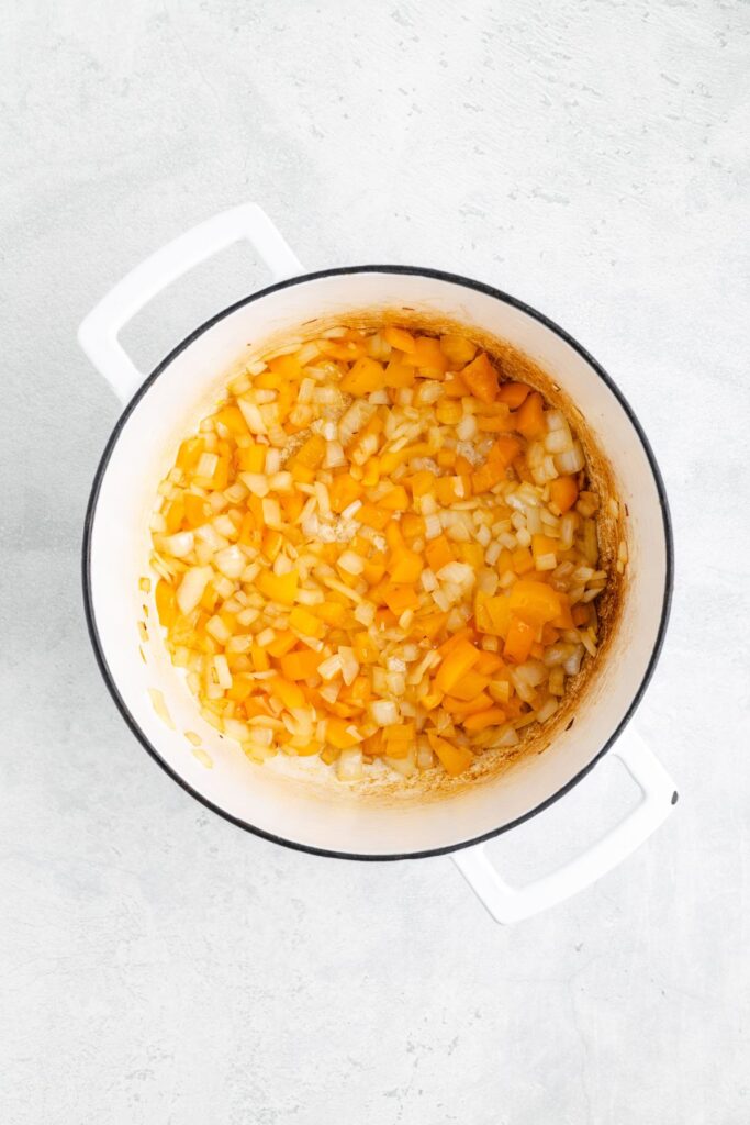 Chopped onions and yellow bell peppers sautéing in a white pot, viewed from above on a light gray surface.