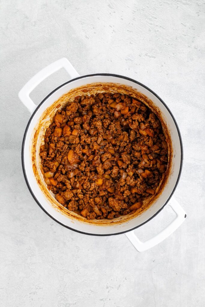 White pot containing cooked ground beef and chopped onions sits on a light gray surface.