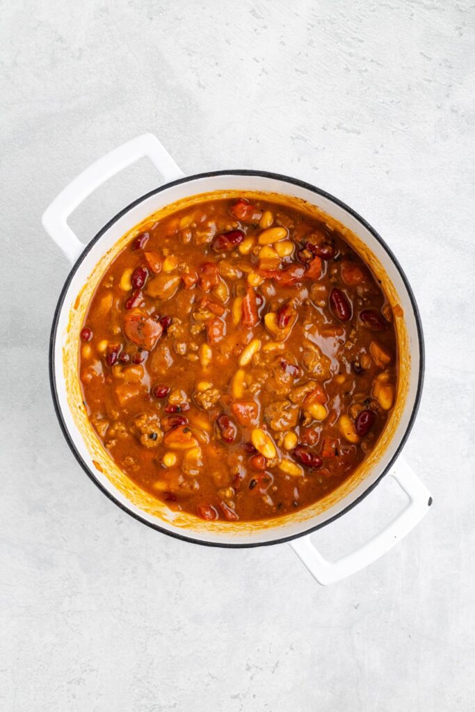 White pot of bean chili with kidney beans, corn, and meat in tomato sauce, on a light gray surface.
