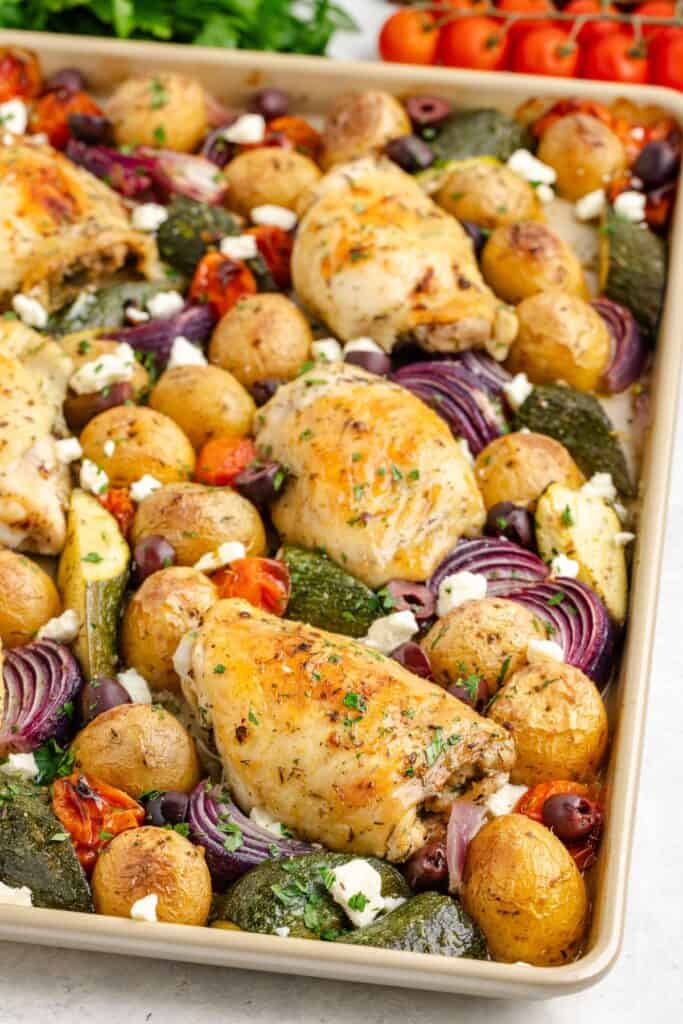 Sheet pan with Greek chicken thighs, potatoes, zucchini, red onion, tomatoes, olives, and feta cheese.