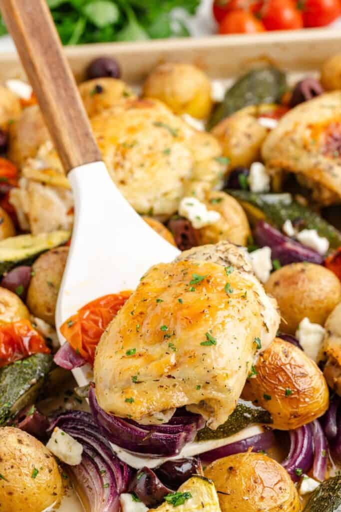 A spatula lifts Oven Roasted Greek Chicken with roasted potatoes, zucchini, tomatoes, olives, and red onions on a sheet pan.