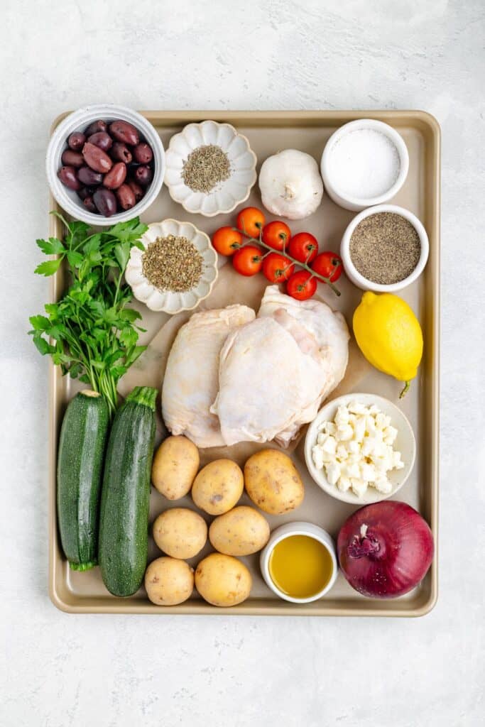 Baking sheet with raw chicken, potatoes, zucchini, tomatoes, red onion, lemon, olives, feta, herbs and spices.