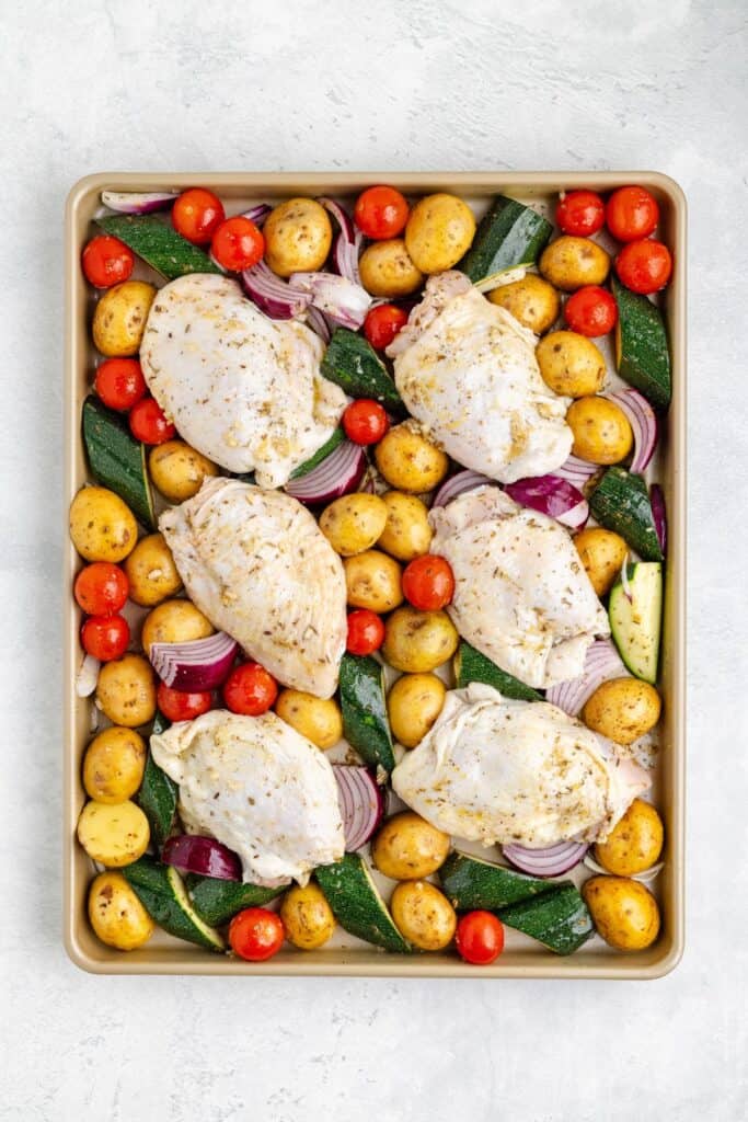 Sheet pan with raw seasoned chicken breasts, cherry tomatoes, baby potatoes, zucchini, and red onion ready to bake.