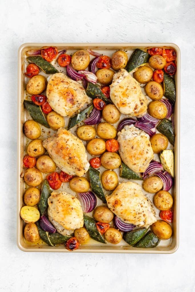 Sheet pan with baked chicken thighs, baby potatoes, zucchini, red onion, and cherry tomatoes arranged together.