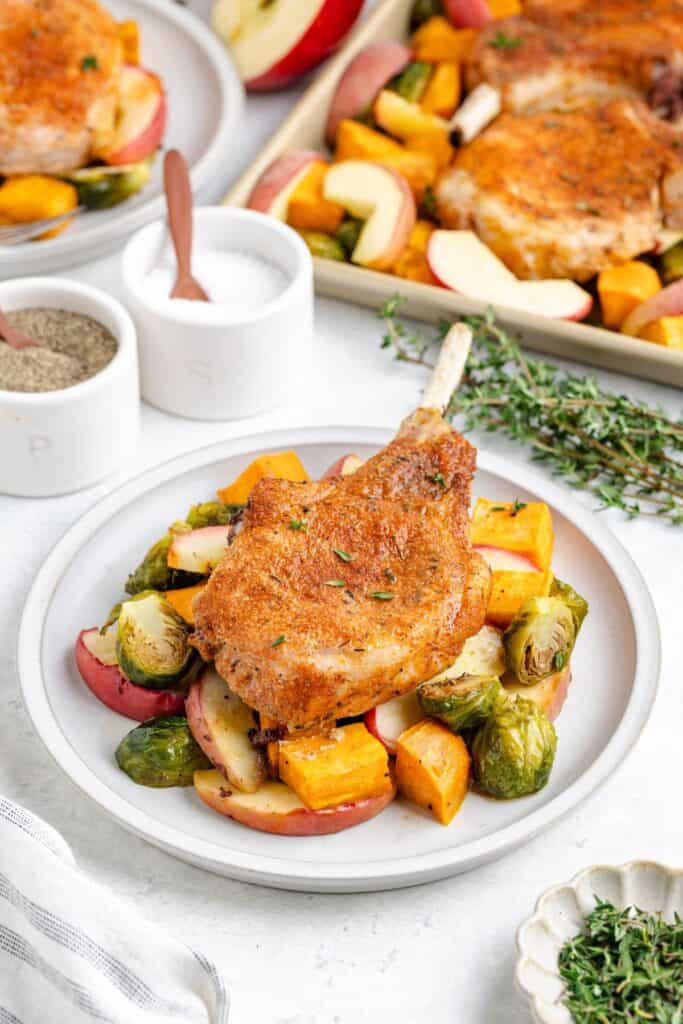 Seasoned baked pork chop atop roasted Brussels sprouts, apple slices, and sweet potatoes on a plate; tray in background.