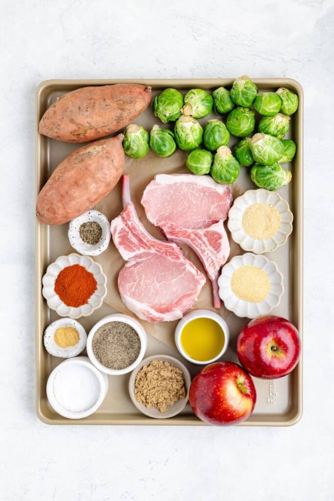 Sheet pan with raw pork chops, sweet potatoes, Brussels sprouts, apples, and assorted spices arranged neatly.