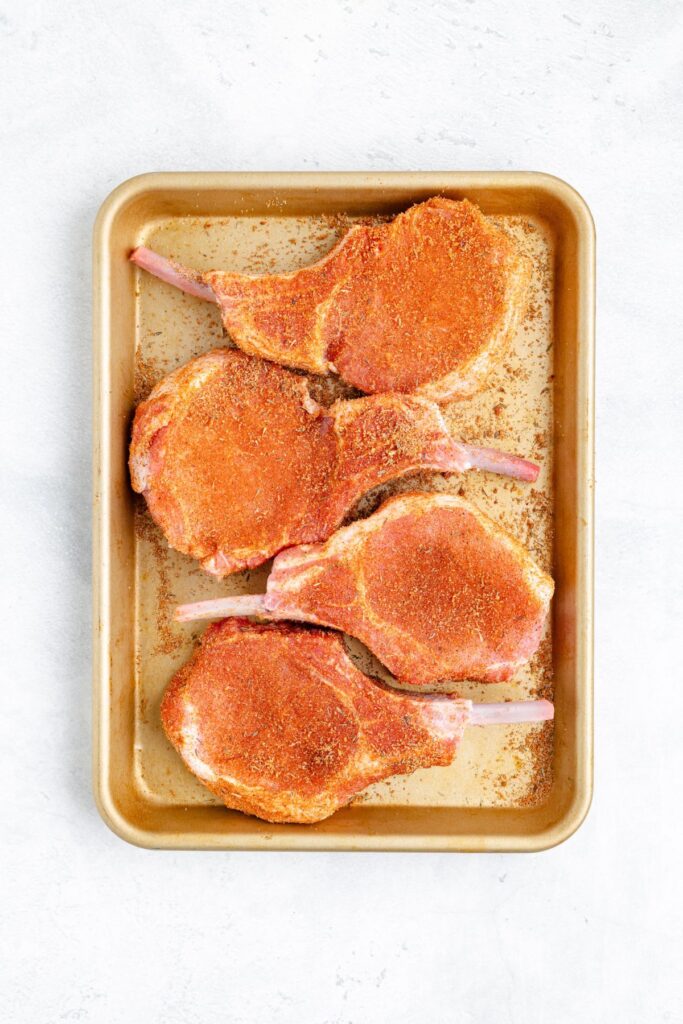 Four uncooked seasoned pork chops on a metal baking sheet, ready to be cooked, with a light background.