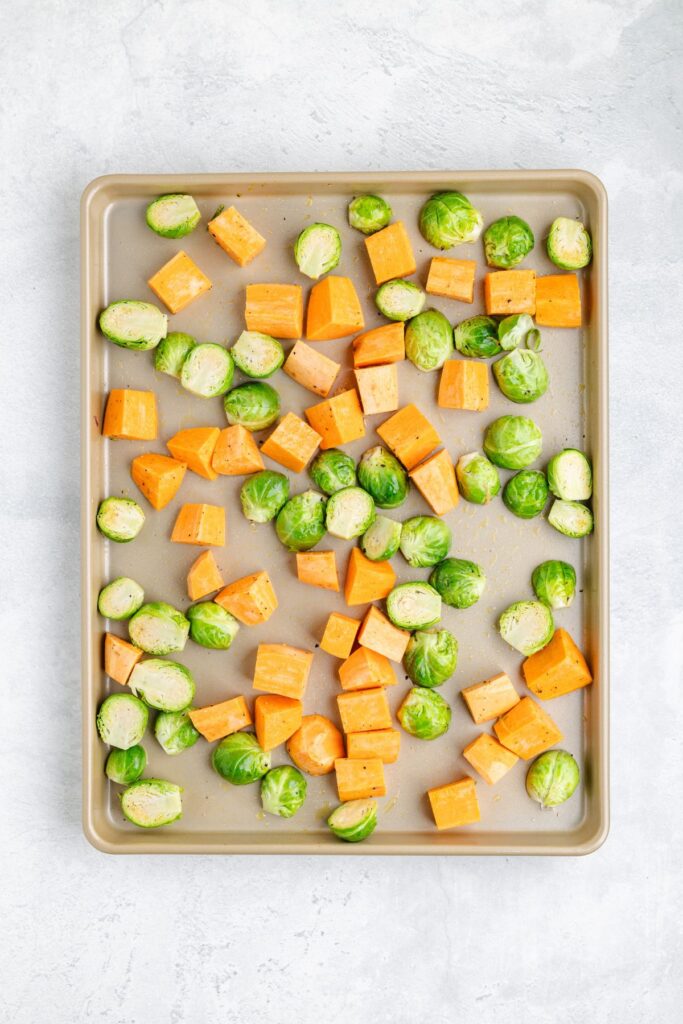 Raw Brussels sprouts halves and sweet potato cubes are spread evenly on a baking sheet against a light background.