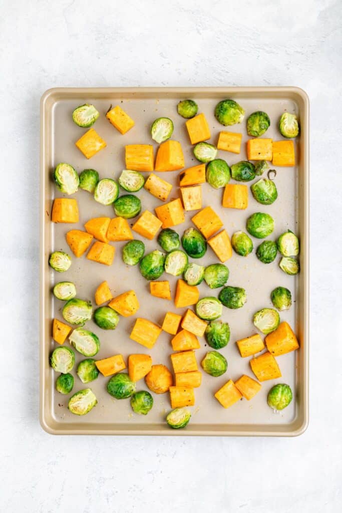 Raw Brussels sprouts halves and sweet potato cubes are evenly arranged on a baking sheet against a light background.