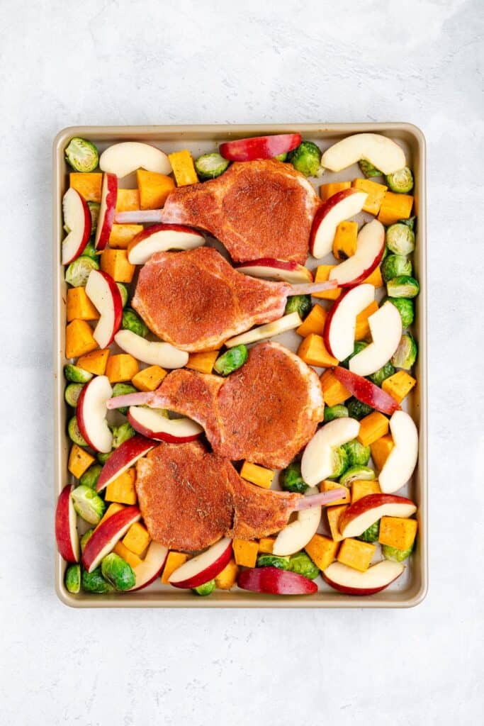 Sheet pan featuring three seasoned pork chops with sliced apples, brussels sprouts, and diced butternut squash.