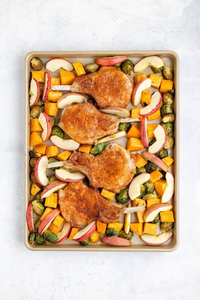 Four seasoned pork chops with sliced apples, brussels sprouts, and cubed squash on a baking sheet ready to roast.