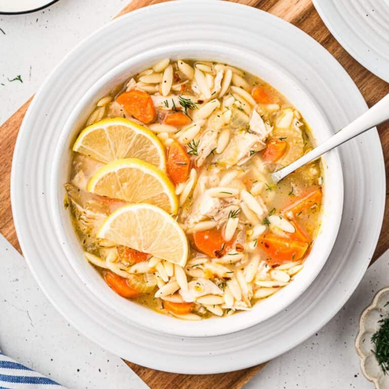 Chicken lemon orzo soup with carrots, herbs, and lemon slices in a bowl on a white plate with a spoon.