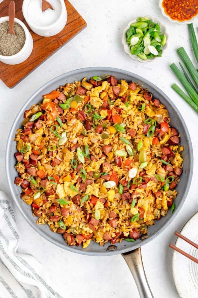A skillet of fried rice with sausage, eggs, veggies, and green onions. Seasonings and toppings surround it.