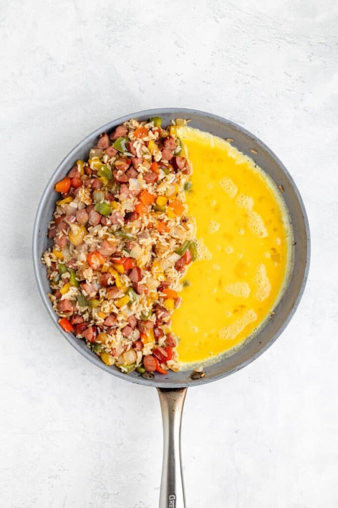 Frying pan with fried rice and veggies on one side, raw beaten eggs on the other, ready to be mixed.