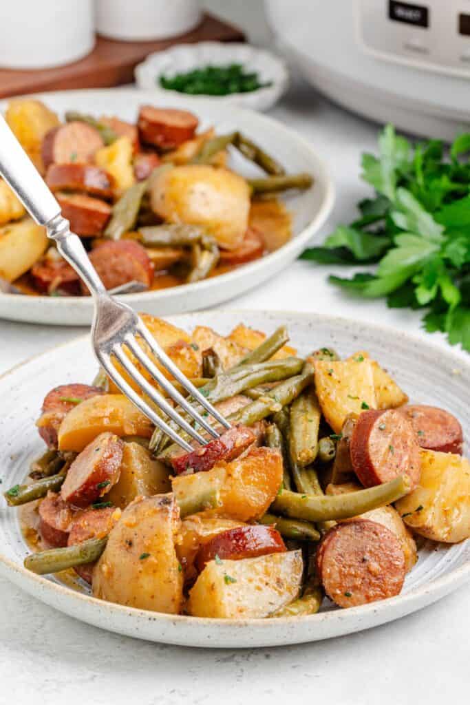 Plate of viral swamp potatoes, sausage, and green beans with a fork in a potato; extra plate and herbs in background.