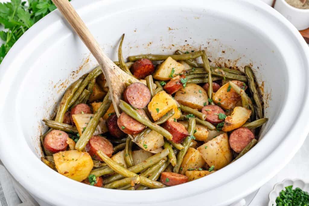 Slow cooker swamp potatoes with green beans and sausage being stirred with a wooden spoon.
