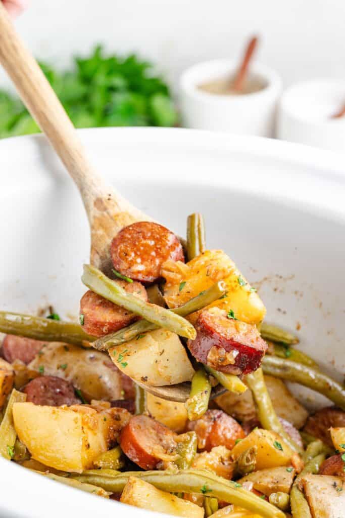 A wooden spoon lifts swamp potatoes from a white slow cooker.