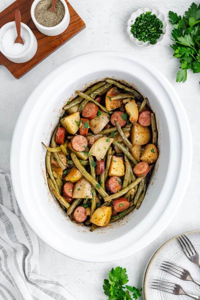 Slow cooker swamp potatoes on a table with plates, seasonings, herbs, and napkin.