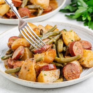 Plate with crockpot swamp potatoes, sausage, and green beans cooked together; a fork lifts a piece of sausage.