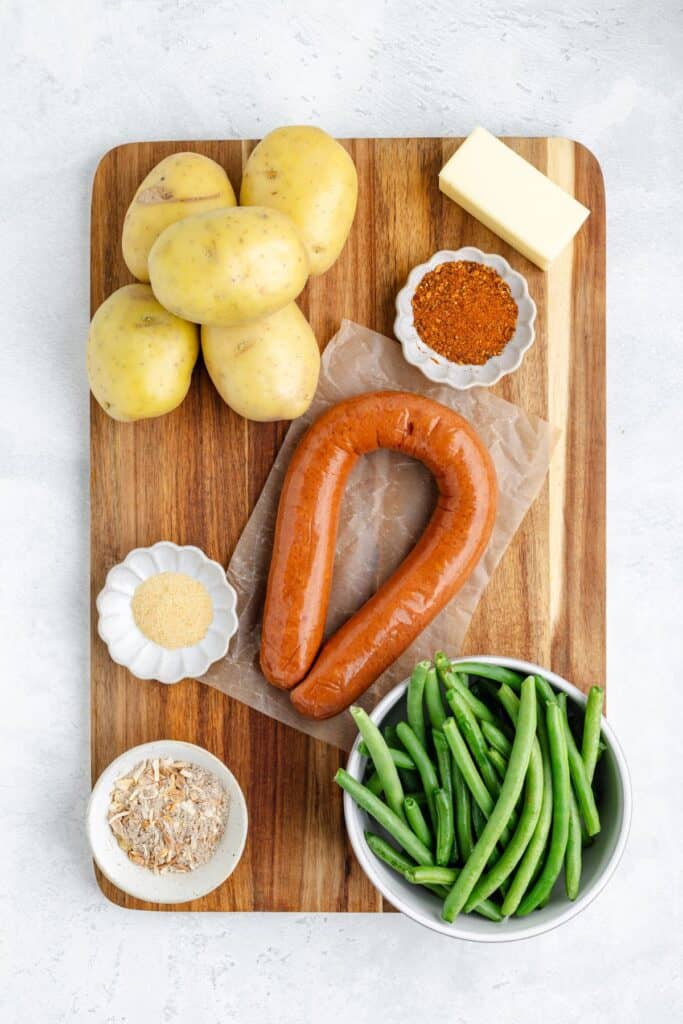 Cutting board with potatoes, butter, sausage ring, green beans, seasoning, and small bowls of spices and dried onions.