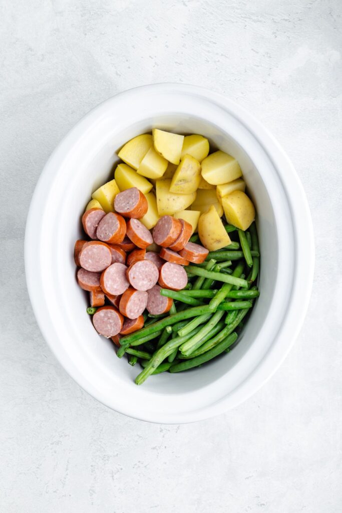 White slow cooker with sliced sausage, chopped potatoes, and green beans arranged in separate sections.