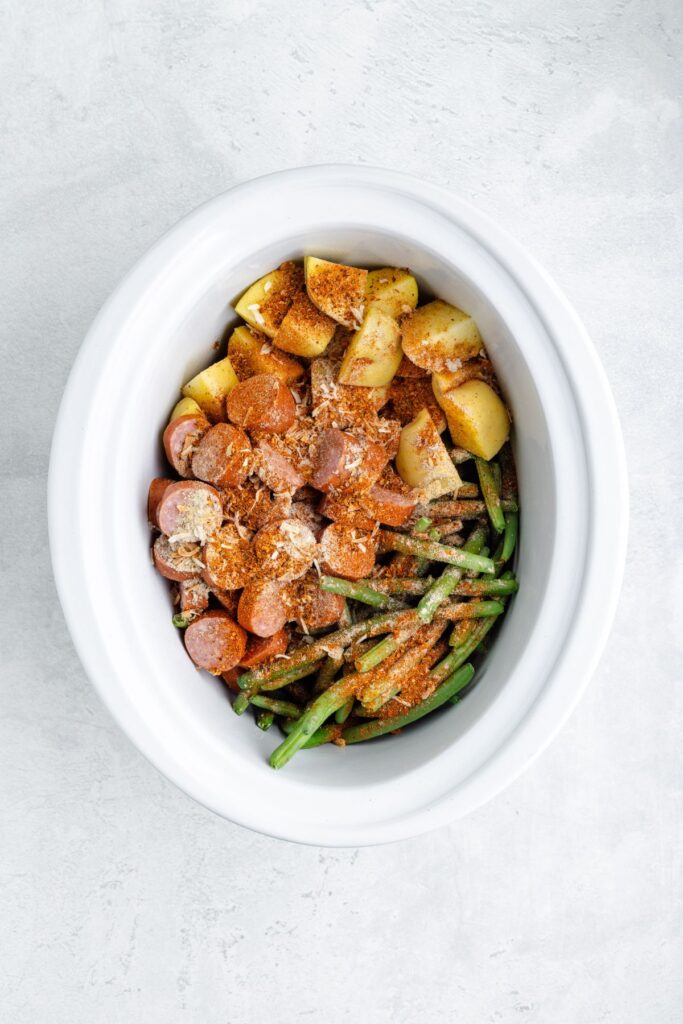 Sliced sausage, green beans, and potatoes with cheese and spices in a white slow cooker on a light surface.