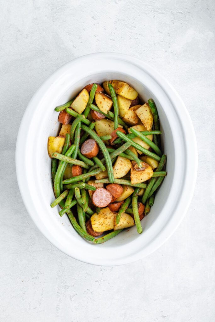 White oval dish with sliced sausage, green beans, and diced potatoes mixed together on a light gray background.