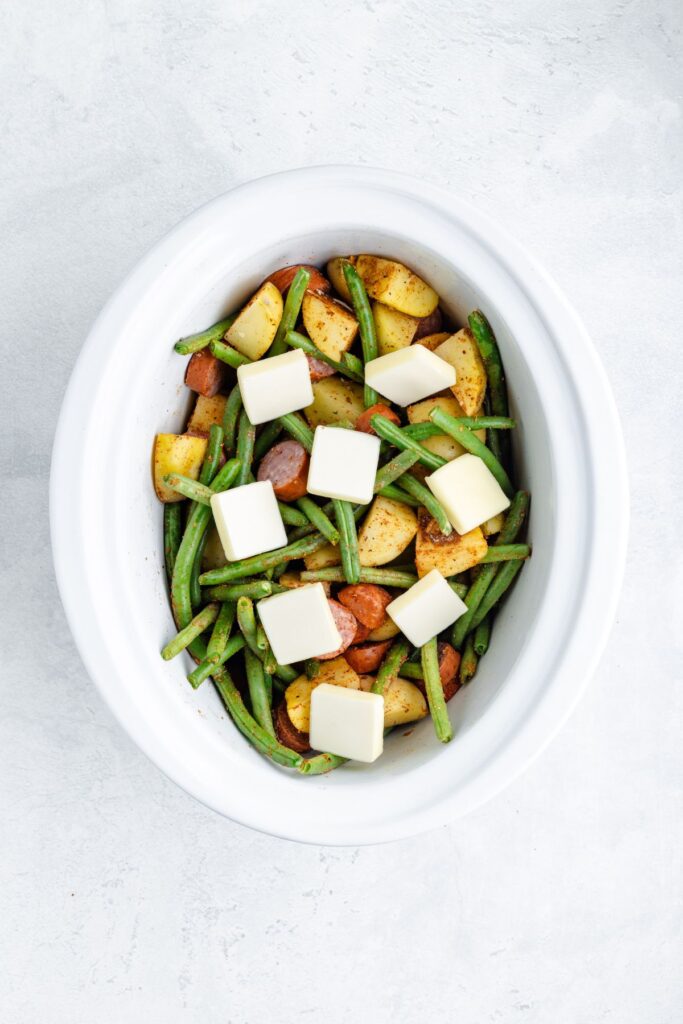 Oval white dish with green beans, sliced potatoes, sausage pieces, and butter cubes on top, ready to cook.