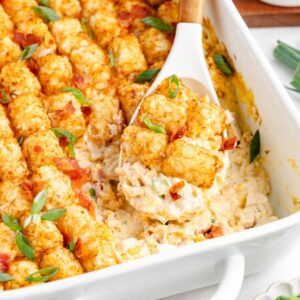 Chicken bacon Ranch tater tot casserole topped with green onions in a white baking dish, a spoon lifting out a serving.
