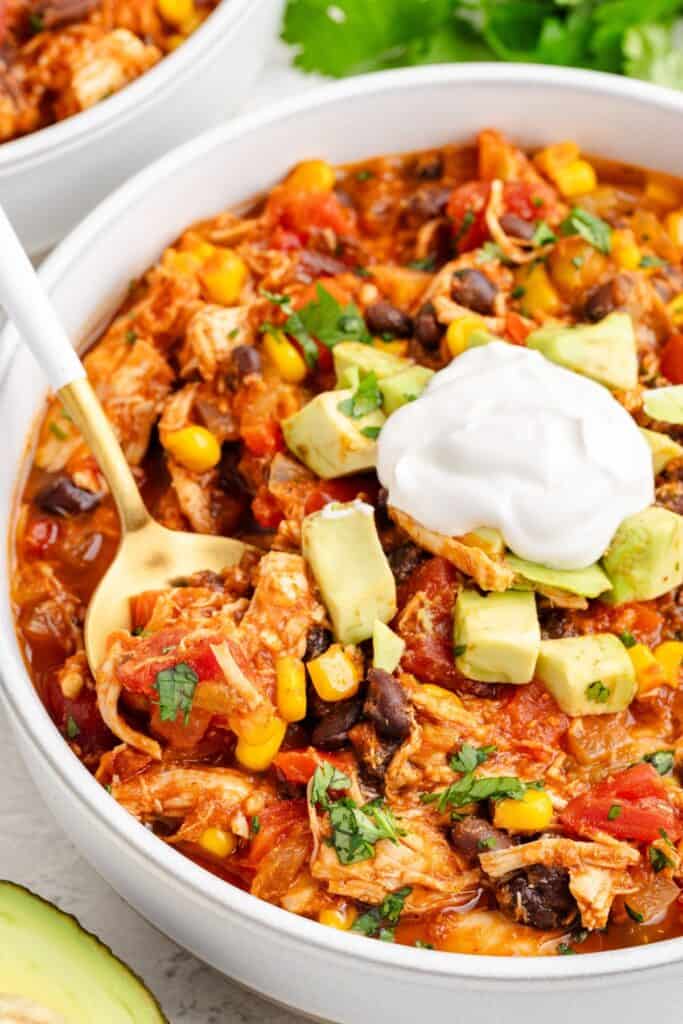 Bowl of red chicken chili topped with avocado, cilantro, sour cream, and a spoon inside.