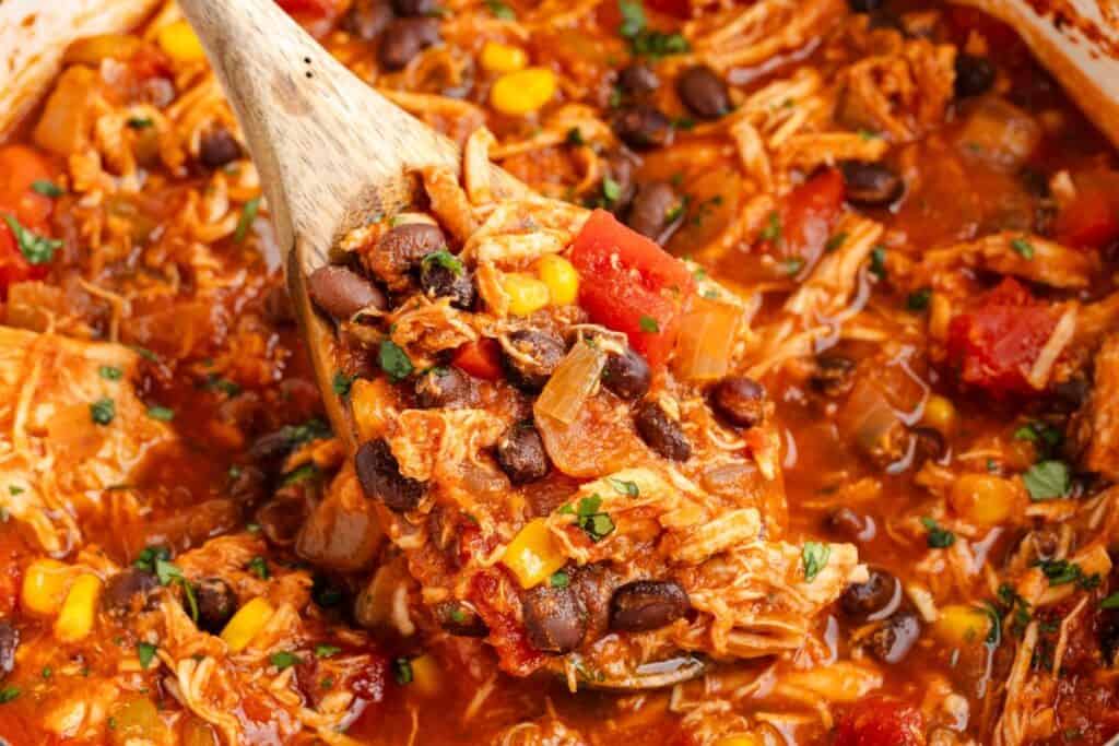 A wooden spoon lifts stovetop red chicken chili with shredded chicken, black beans, corn, tomatoes, and onions in red sauce.