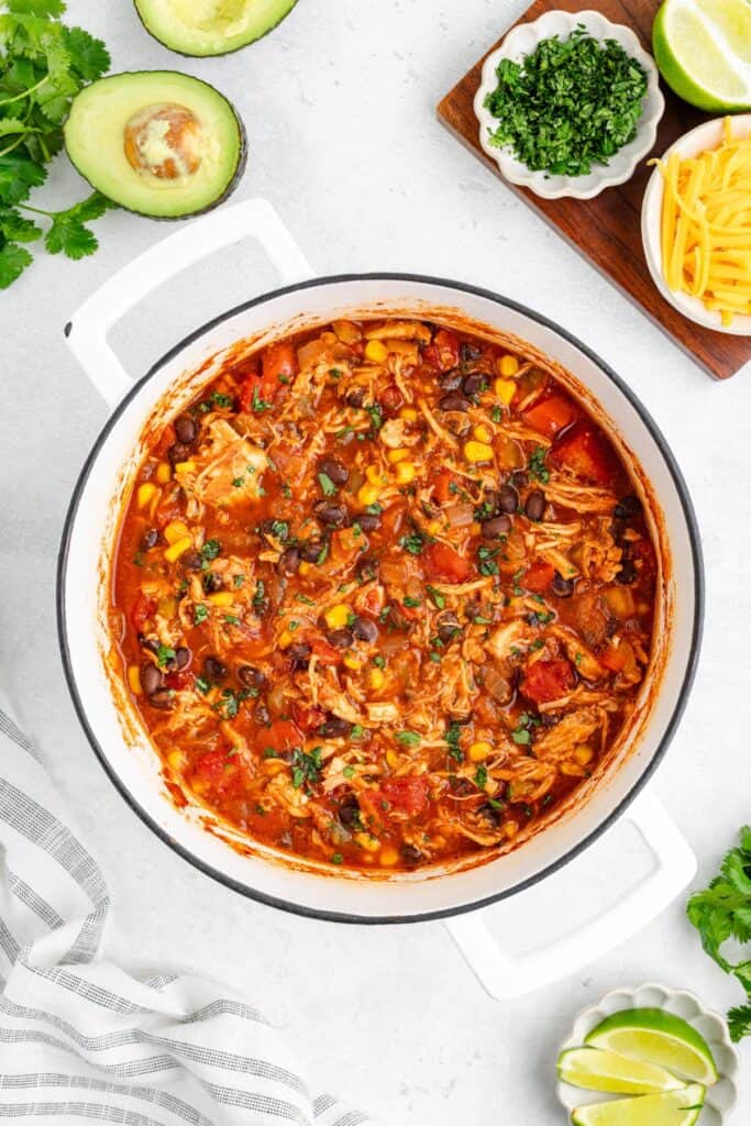 A pot of chicken chili with beans, corn, and tomatoes on white surface, surrounded by taco toppings.