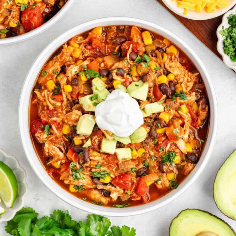 Bowl of chicken red chili with black beans, corn, avocado, sour cream, herbs; lime wedges and avocado on side.
