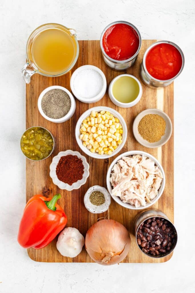 Neatly arranged on a wooden board: broth, canned tomatoes, spices, corn, green chiles, chicken, pepper, beans.