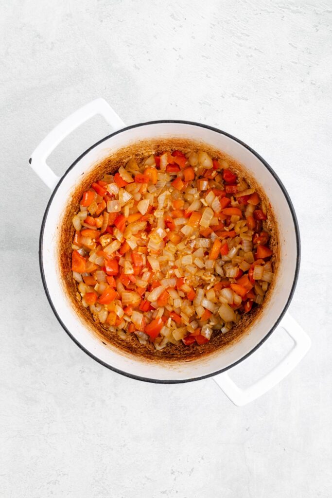 Chopped onions and red bell peppers sauté in a white Dutch oven on a light countertop.