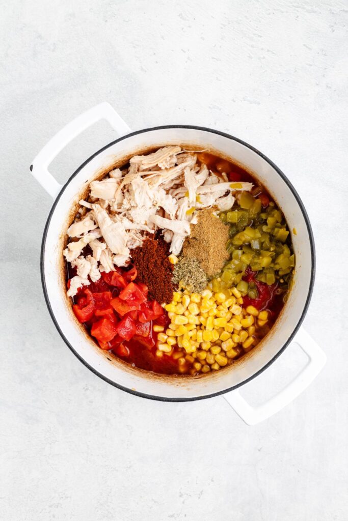 White pot filled with shredded chicken, corn, tomatoes, green chilies, and spices on a light-colored surface.