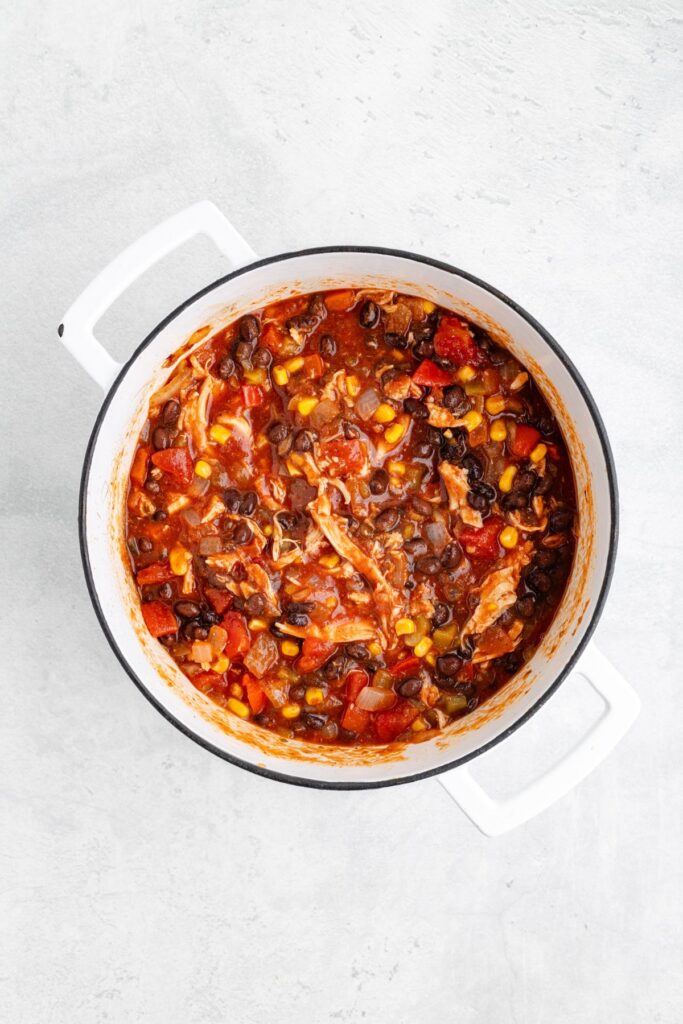 White pot with shredded chicken, black beans, corn, and diced tomatoes in red sauce, top-down view.