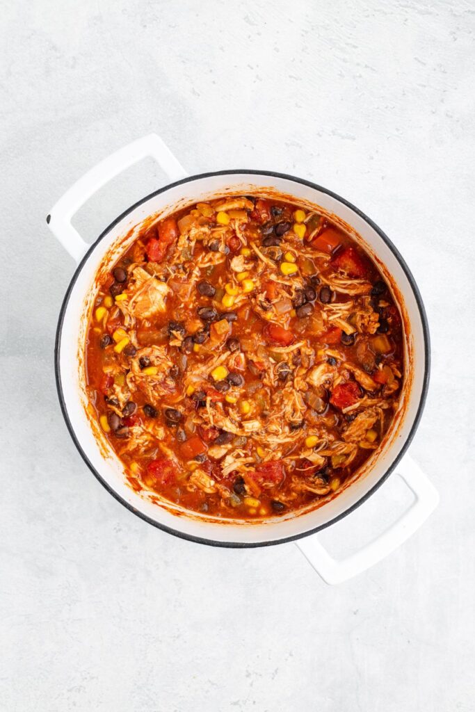 White pot with shredded chicken, black beans, corn, and tomatoes in red sauce, viewed from above.